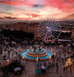 Weihnachtsmarkt mit beleuchtetem Riesenrad und Tritonenbrunnen bei Sonnenuntergang