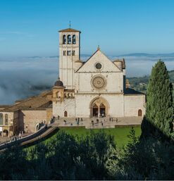 Frontale Ansicht der Basilika San Francesco in Assisi, Italien, bei morgendlichem Sonnenlicht mit Blick auf das Tal und leichtem Nebel im Hintergrund