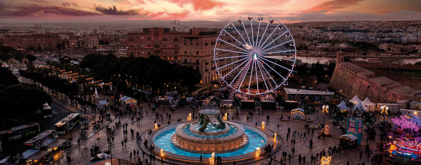 Abendliche Szene mit beleuchtetem Riesenrad, Tritonenbrunnen und Weihnachtsmarkt in Maltas Hauptstadt